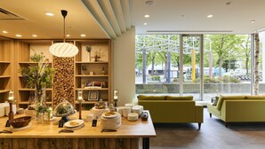 A modern interior with a wooden reception desk, shelves with decorative items, a green sofa, and large windows offering a view of the outdoors.