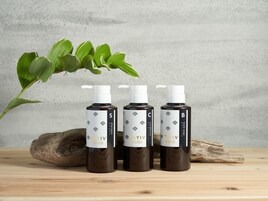 Tres botellas de champú con dosificadores blancos y etiquetas, sobre una superficie de madera con una planta y un trozo de madera al fondo.