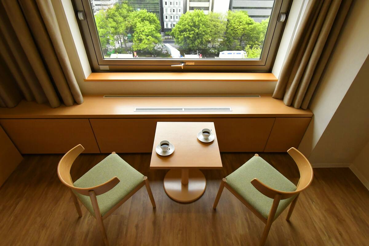 A room with a window seat, two chairs, a small table, and a view of trees and buildings outside.