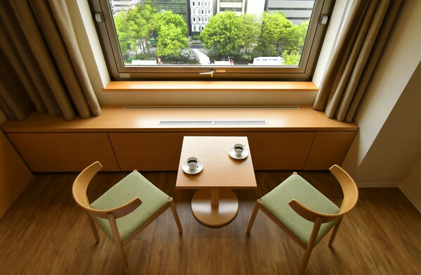Una habitación con un asiento junto a la ventana, dos sillas, una mesita y vistas a árboles y edificios.