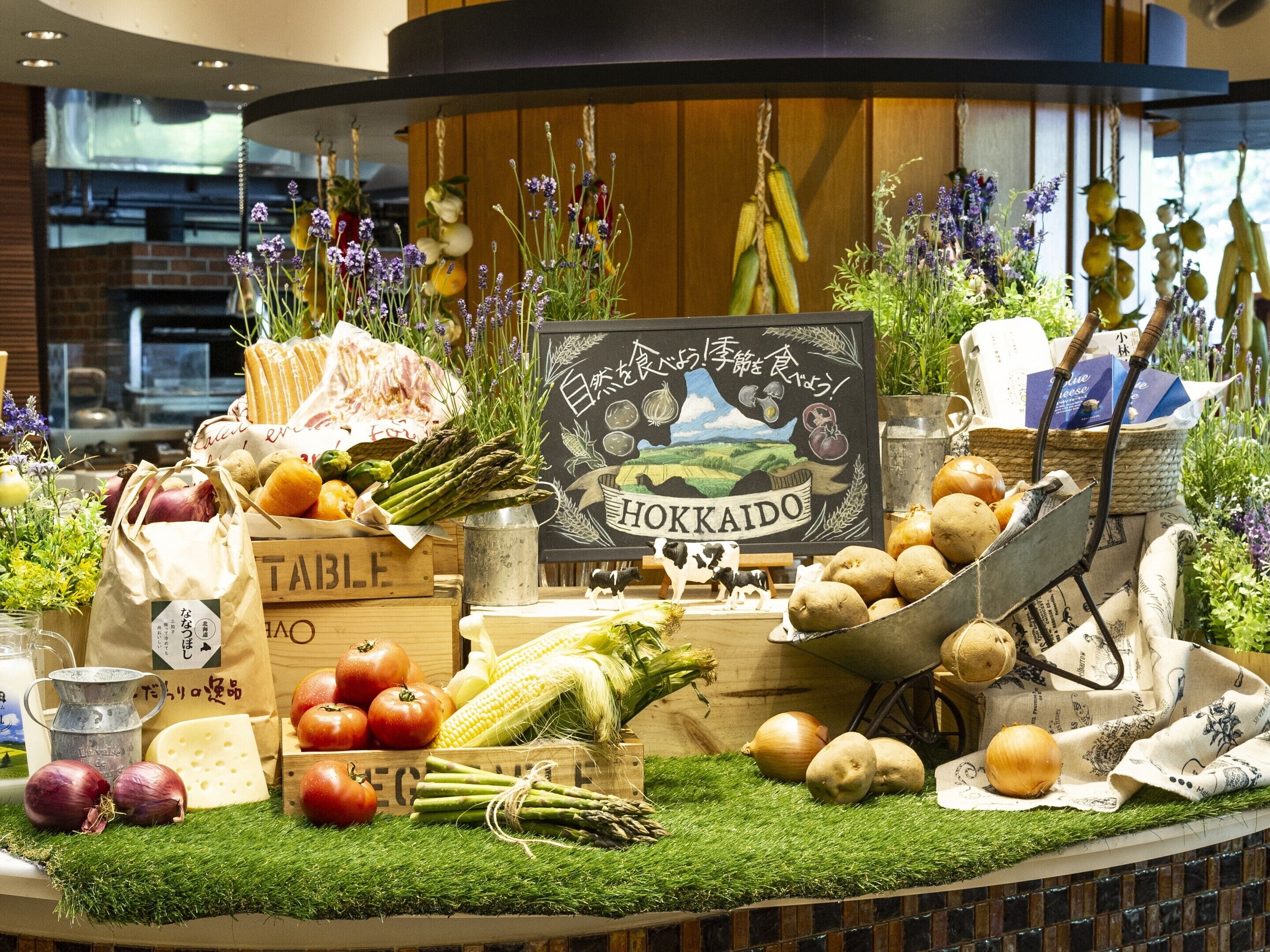 Une présentation de légumes frais et de produits laitiers, notamment des tomates, des asperges et du fromage, disposés sur une table en bois, avec un panneau noir indiquant « HOKKAIDO ».