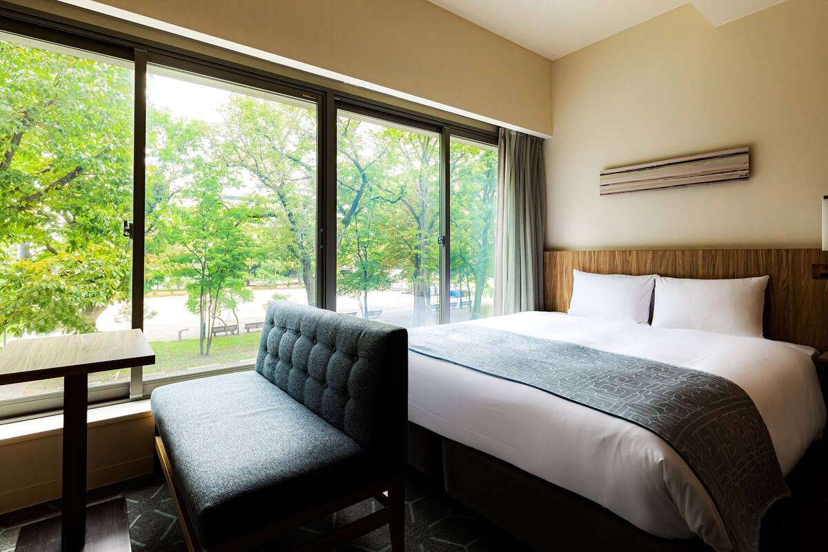 A hotel room with a large bed, a bench, a table, and a window with a view of trees.