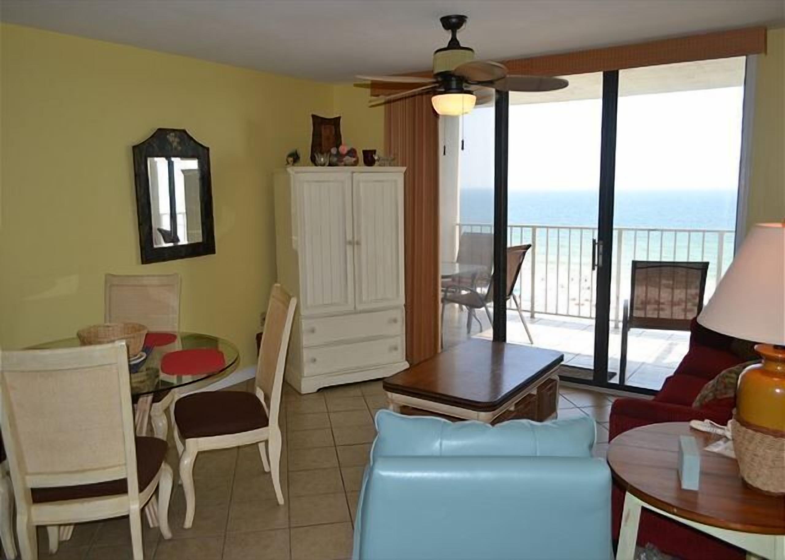 Una habitación con una mesa de comedor, sillas, un sofá, un ventilador de techo, un armario y vistas al océano a través de un ventanal corredera.