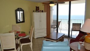 A room with a dining table, chairs, a sofa, a ceiling fan, a cabinet, and a view of the ocean through a sliding glass door. - Sunswept Condominiums (Orange Beach)