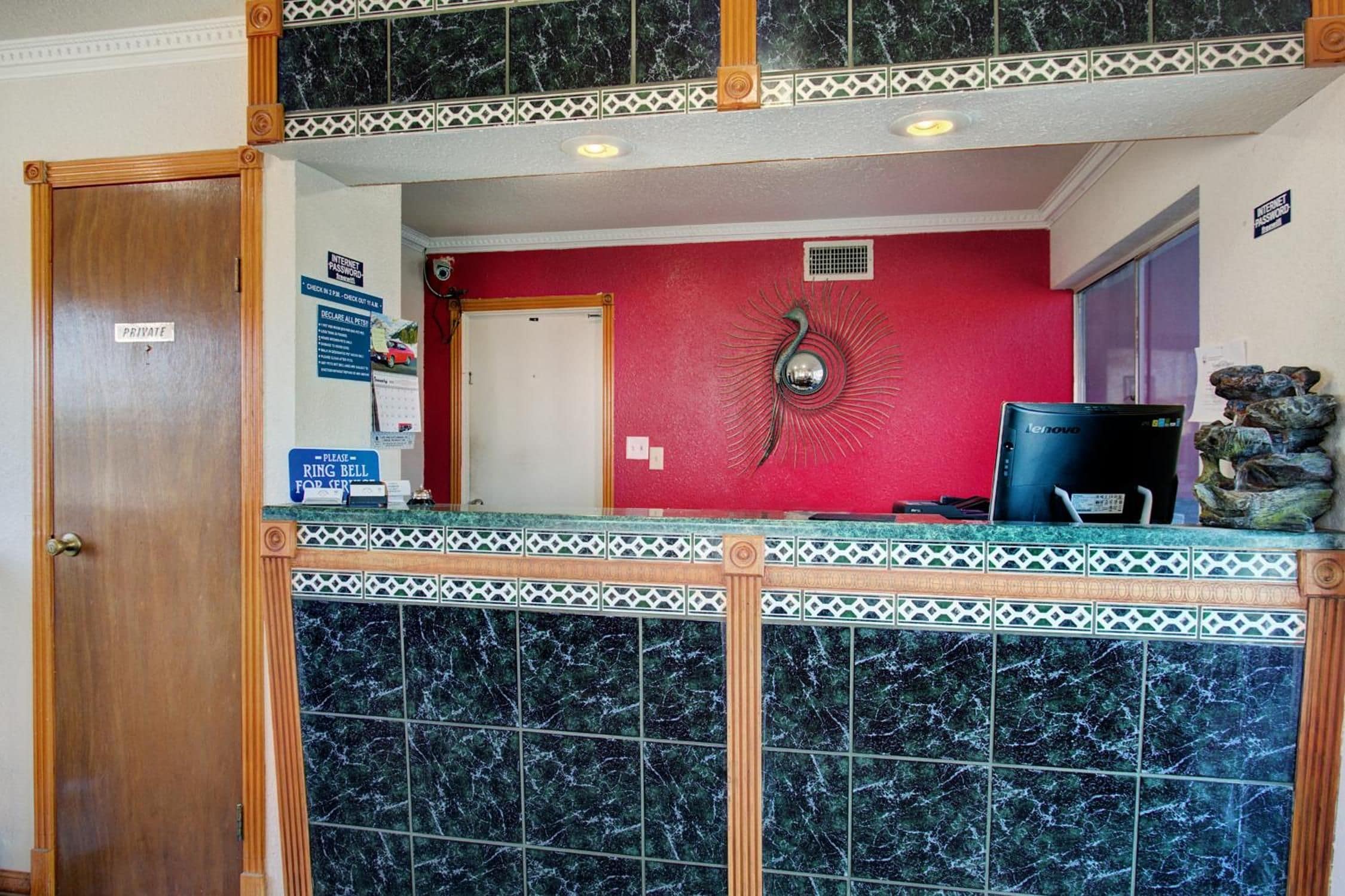 A reception area with a marble counter, a red wall with a decorative metal piece, a Lenovo computer, and a private door.