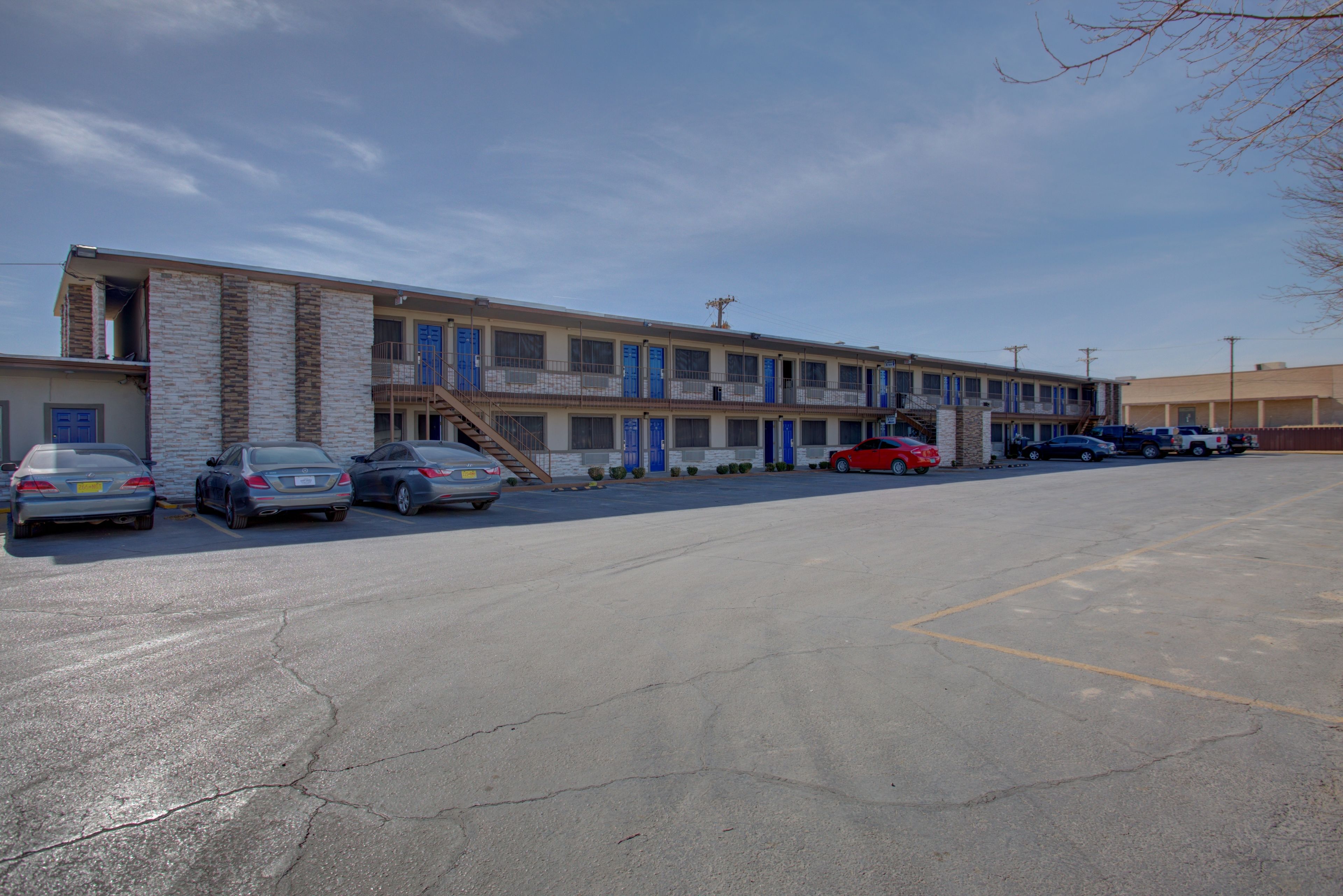 Un motel doté d’un parking, où plusieurs voitures sont garées, et d’un bâtiment à la façade en briques et aux portes bleues.