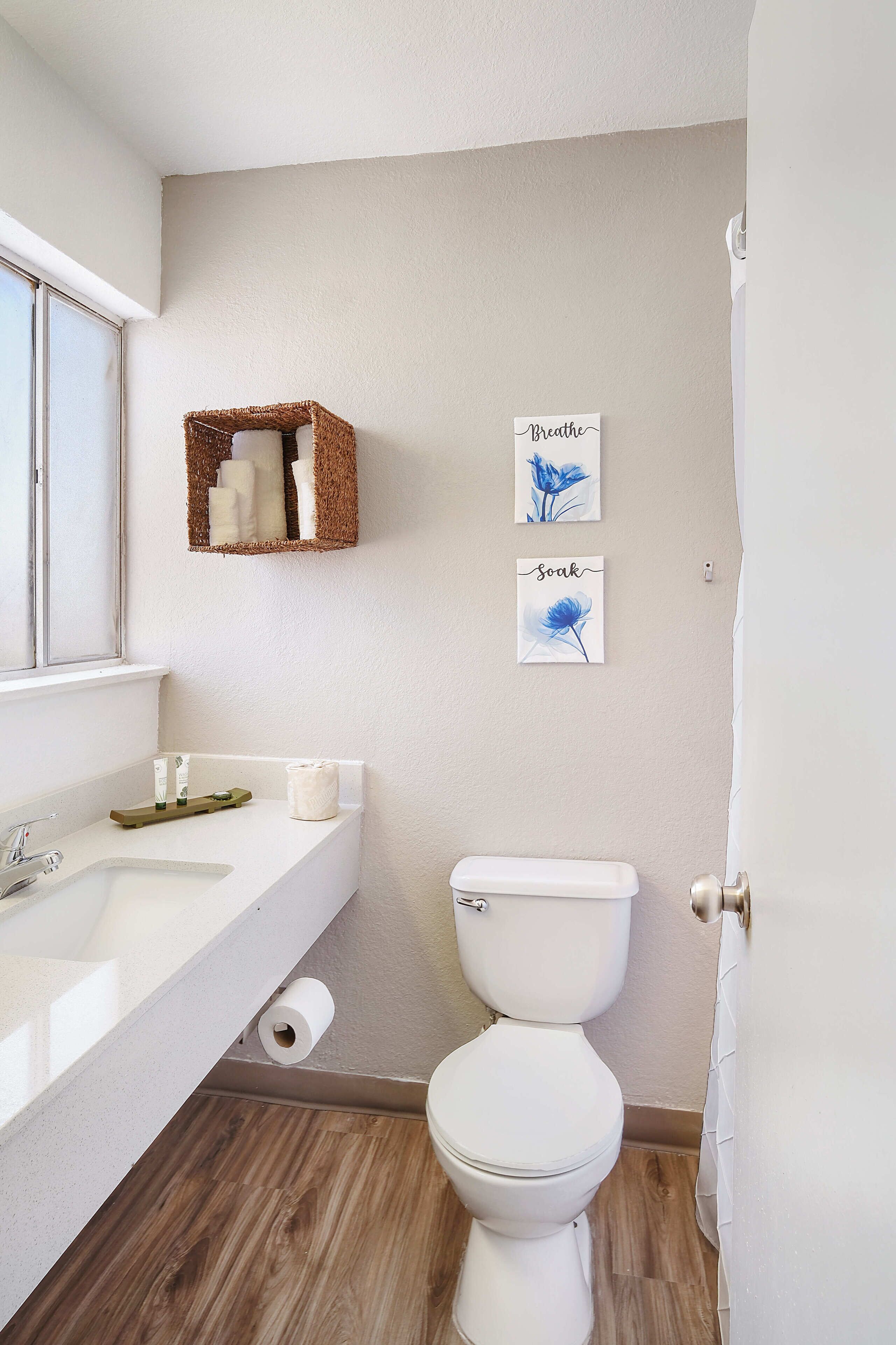 A bathroom with a white sink, toilet, and wooden flooring. There are two wall-mounted baskets and two framed pictures.