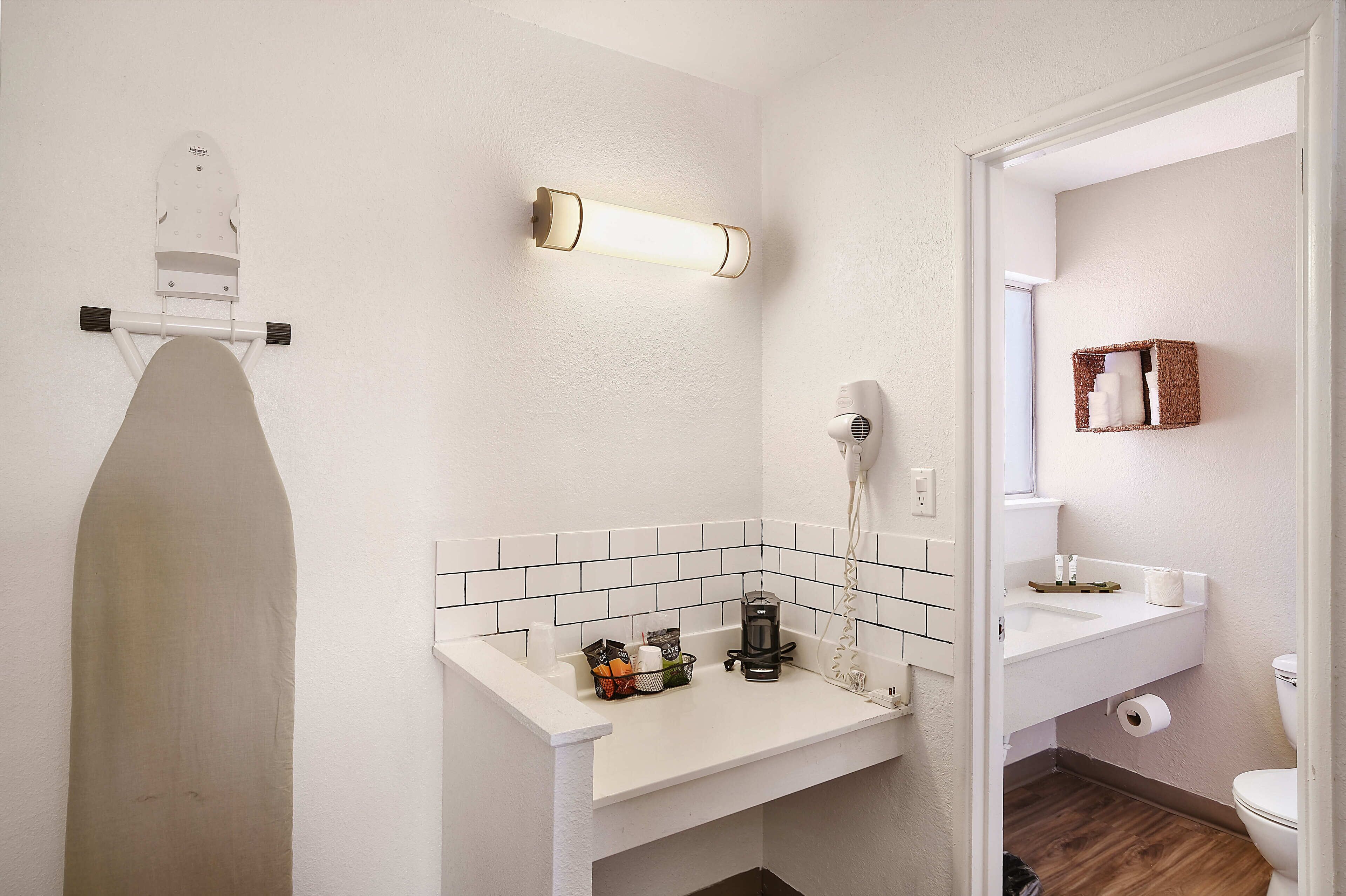 Un baño con lavamanos blanco, secador de pelo, espejo y estante pequeño con artículos de aseo.