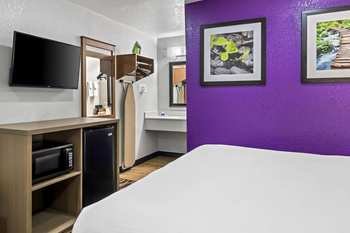 Ein Hotelzimmer mit einem Bett, einem Flachbildfernseher, einer Mikrowelle, einem Kühlschrank, einem Holzregal, einem Badezimmer mit Spiegel und zwei gerahmten Bildern an der Wand.