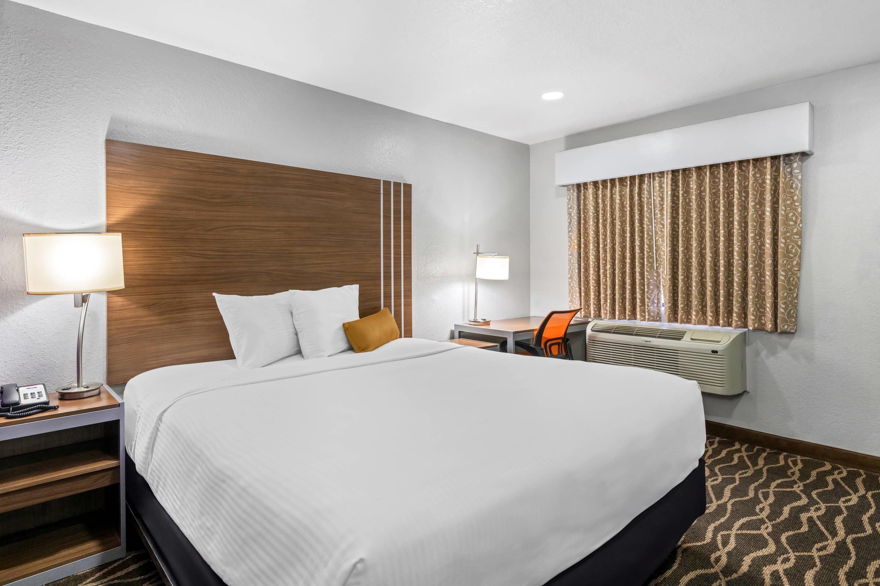 A hotel room with a large bed, bedside tables with lamps, a desk, and an air conditioning unit.