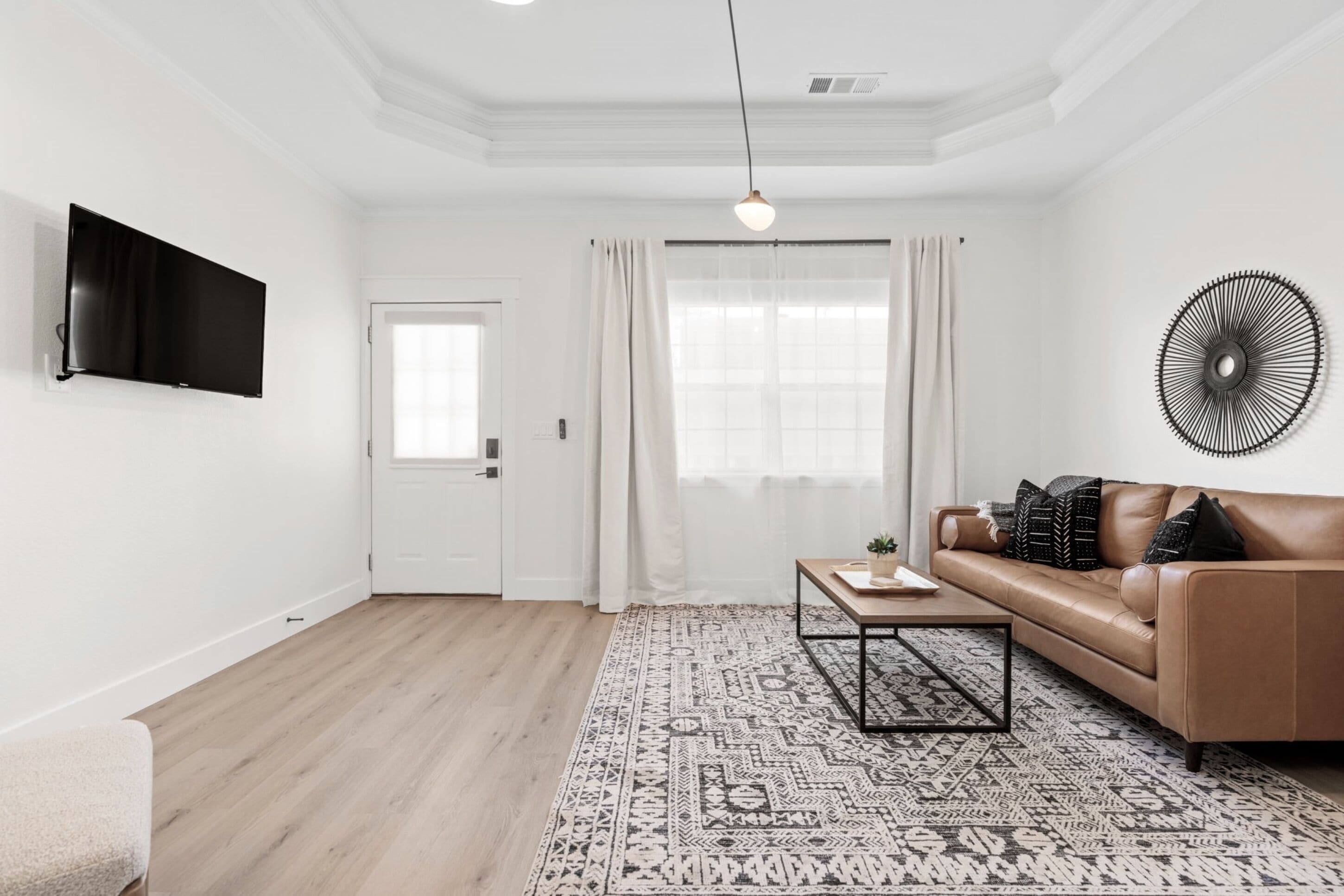 A modern living room with a brown leather sofa, a patterned rug, a coffee table, a flat-screen TV mounted on the wall, and a decorative wall piece.
