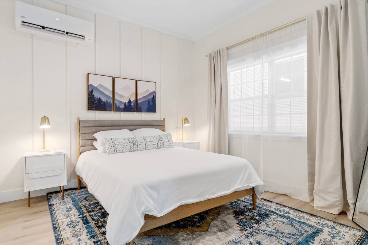 Quarto com cama, mesas de cabeceira, tapete, janela com cortinas e obra de arte na parede.