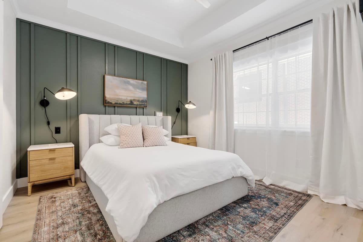Quarto com uma cama grande, duas mesas de cabeceira, um quadro na parede e uma janela com cortinas leves.