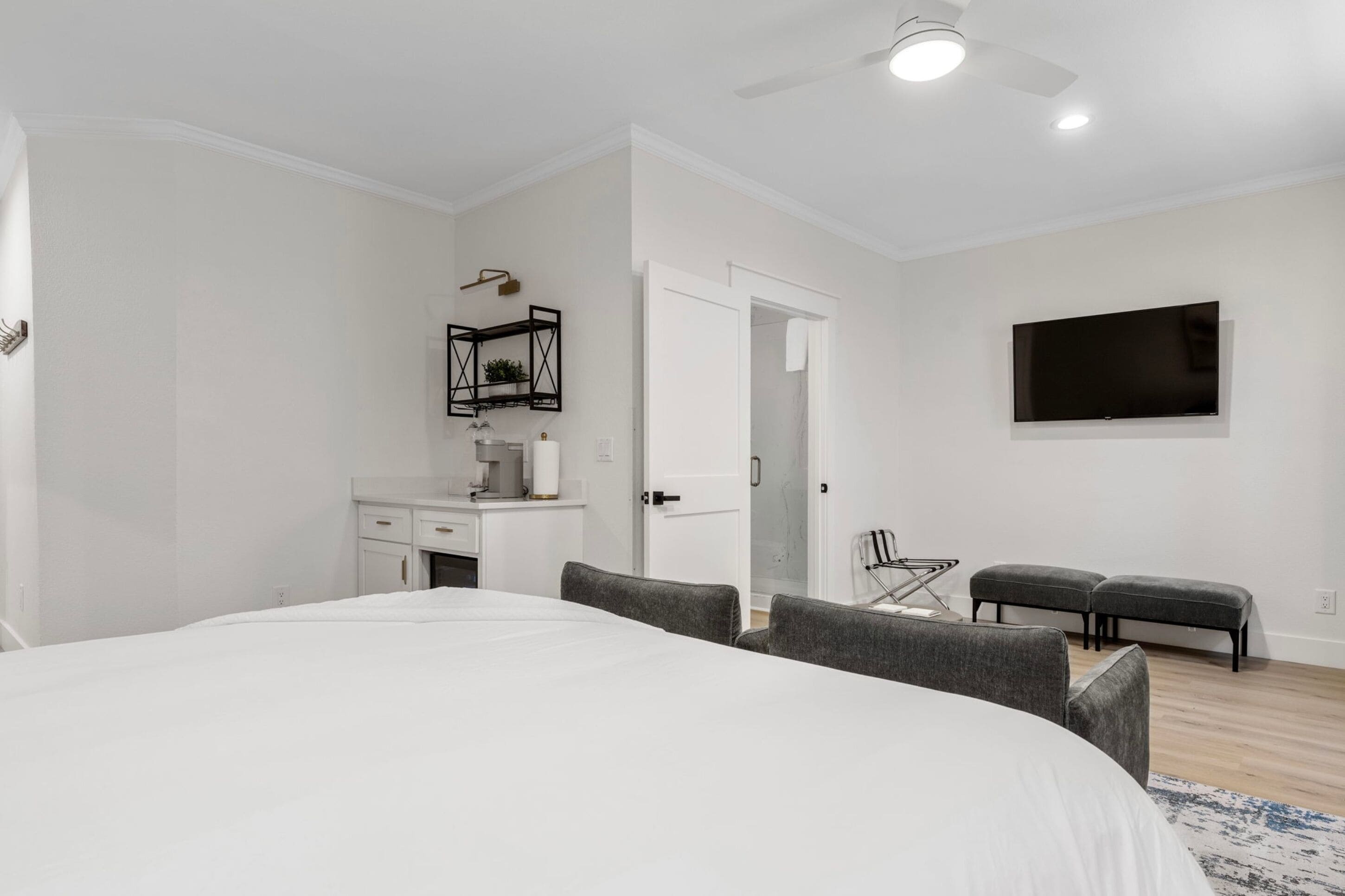 A bedroom with a large bed, a desk with a chair, a TV mounted on the wall, and a door leading to another room.