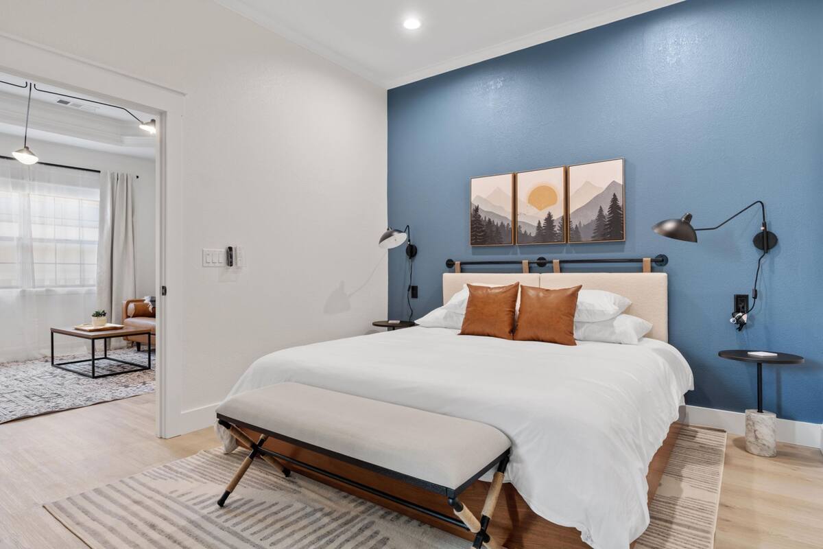 Quarto moderno com uma cama grande, um banco, uma escrivaninha e uma cadeira. Há duas luminárias fixadas na parede e um quadro sobre a cama.