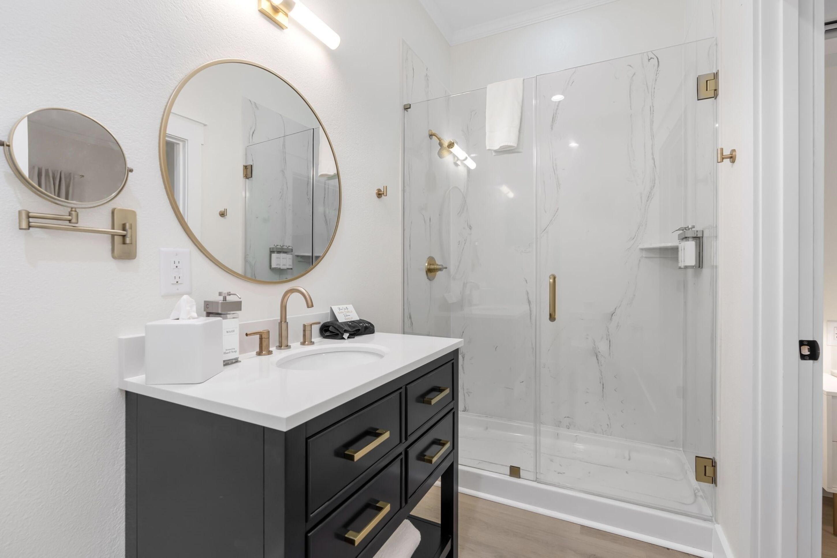 A modern bathroom with a white marble shower, a round mirror, and a dark vanity with gold handles.