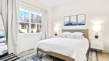 A modern bedroom with a large bed, a nightstand, a lamp, and a view of the outdoors through a window.
