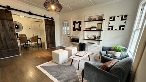 A modern living room with a sofa, ottoman, coffee table, and a large rug. There is a sliding barn door, a dining area with chairs, and a fireplace with decorative items. - 27 On Park (Dahlonega)