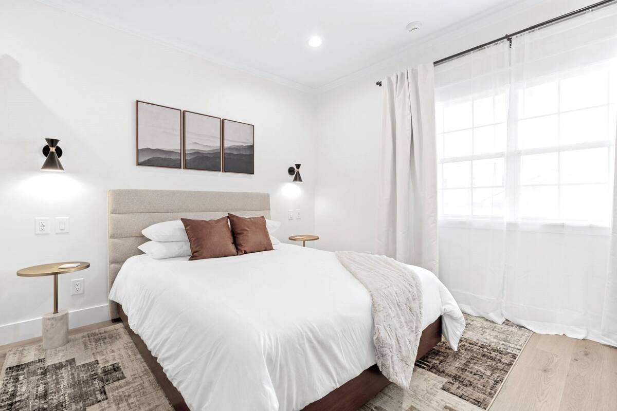 Quarto moderno com uma cama grande, duas mesas de cabeceira, um painel de cabeceira e uma janela com cortinas leves.