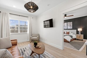 Una habitación de hotel moderna con un ventanal grande, un sofá, una mesa de centro, un televisor, una cama y una mesita de noche con una lámpara.
