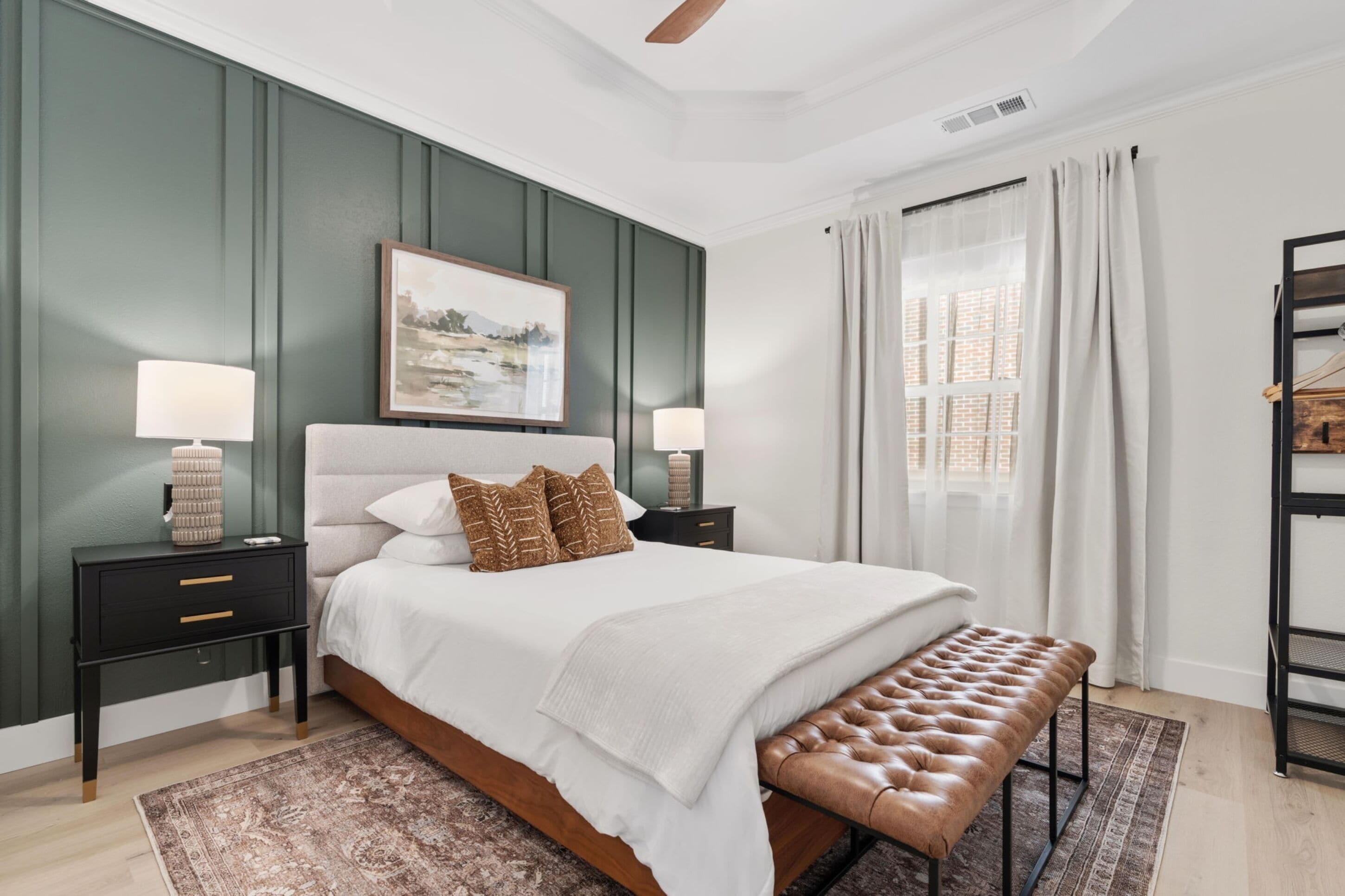 Une chambre à coucher avec un grand lit, deux tables de chevet, un banc, un tableau et une fenêtre avec des rideaux.