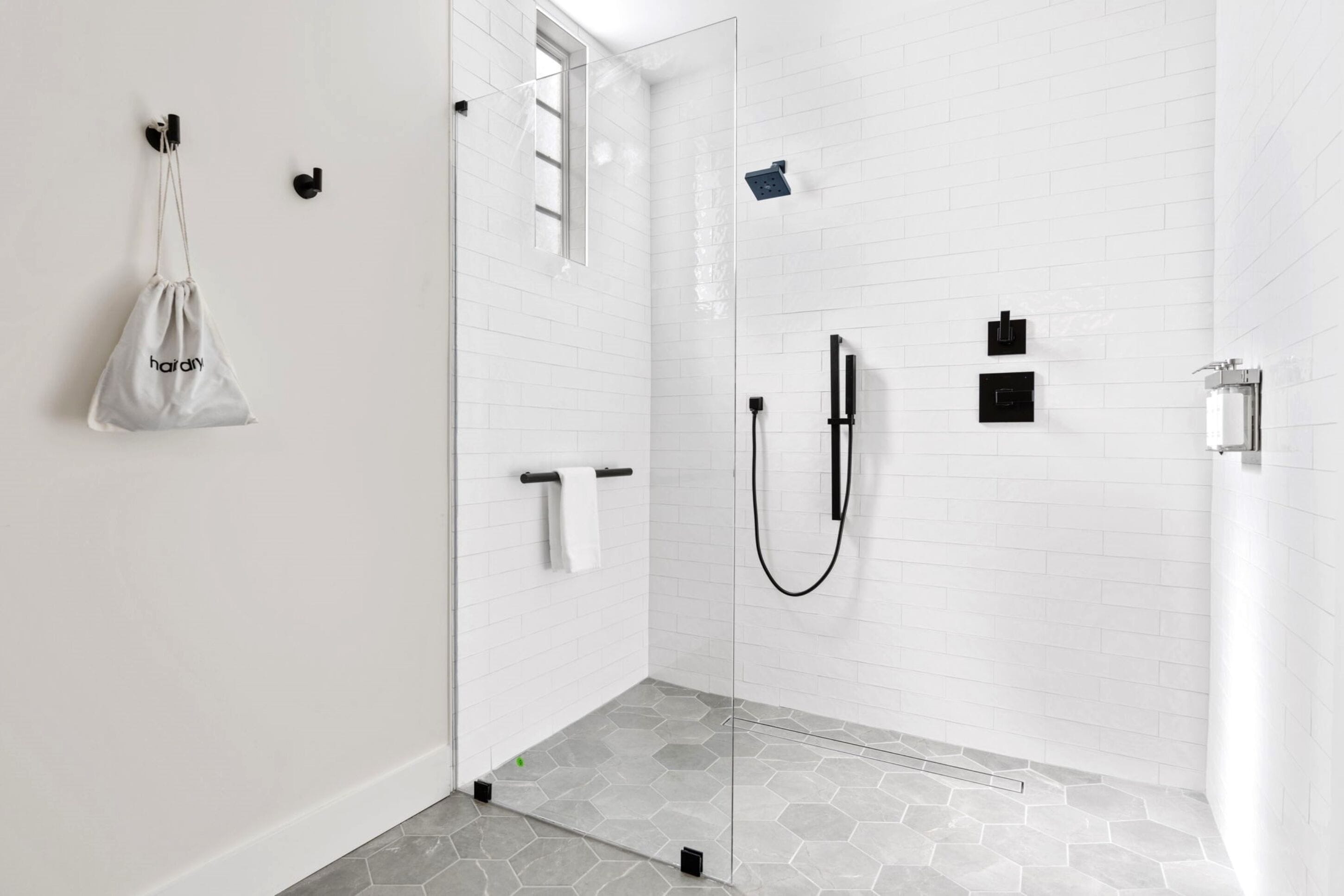 Un baño moderno con azulejos blancos tipo metro, cabina de ducha de vidrio, toallero y una bolsa colgante con la etiqueta 'hairdry'.