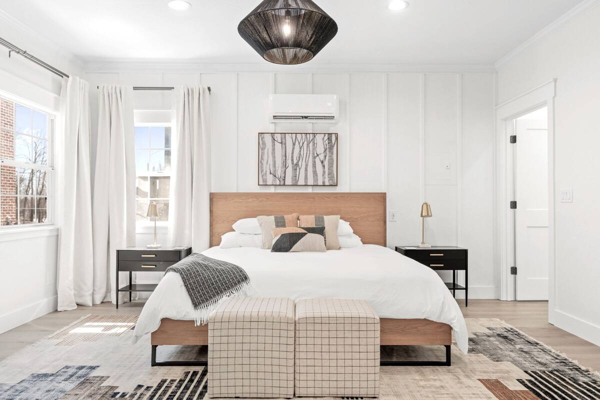 Quarto com cama grande, mesas de cabeceira, luminária pendente e janela com cortinas.