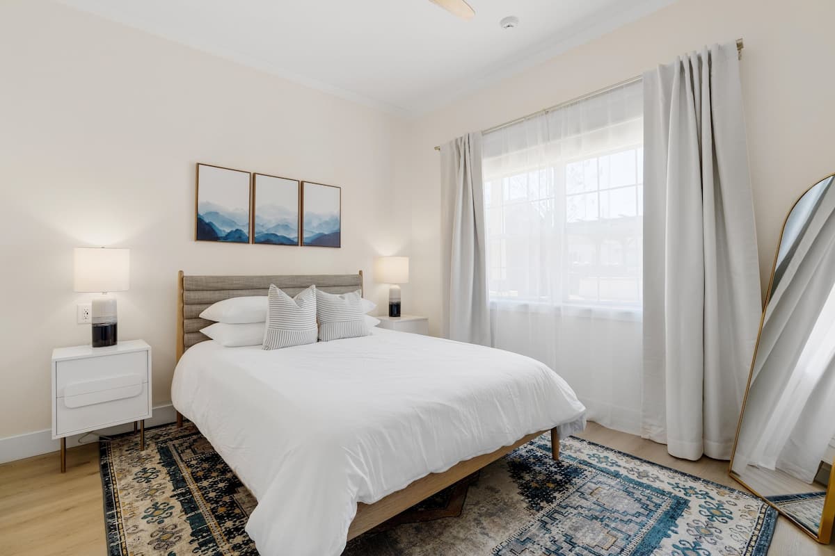 Quarto bem arrumado com uma cama grande, duas mesas de cabeceira com luminárias, um tapete e uma janela ampla com cortinas.