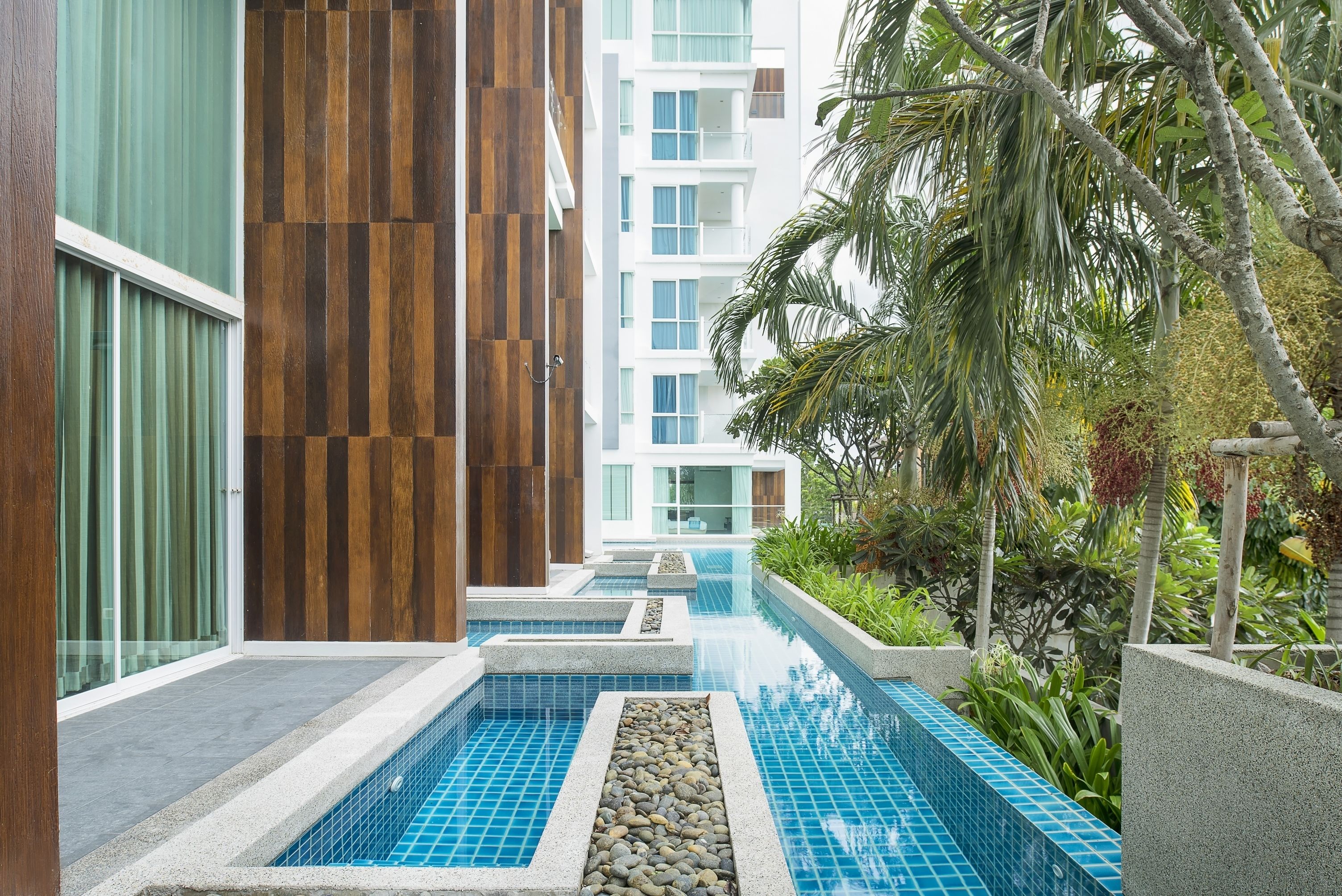 A modern building with a pool area, featuring a stone pathway leading to the pool, surrounded by greenery and a multi-story structure in the background.