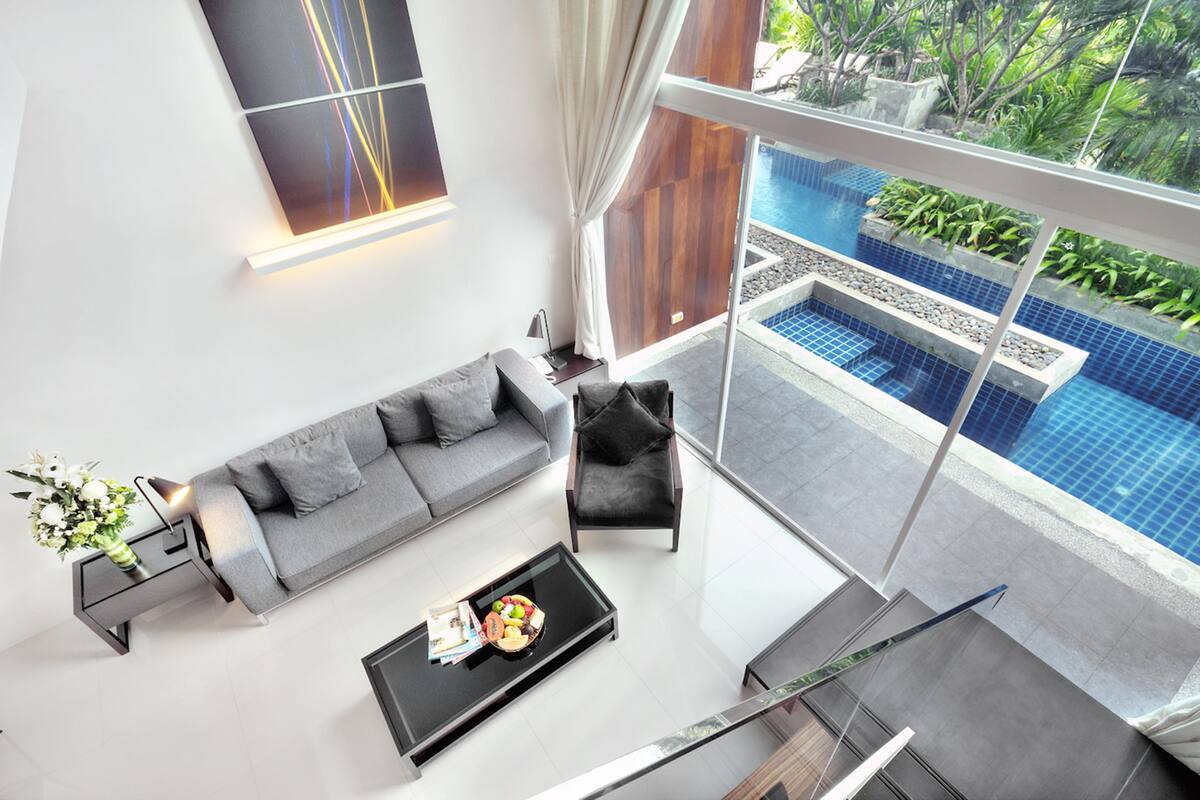 Una sala de estar moderna con un sofá gris, un sillón negro y una mesa de centro de vidrio con frutas y revistas. Un ventanal ofrece vistas a una piscina y abundante vegetación.