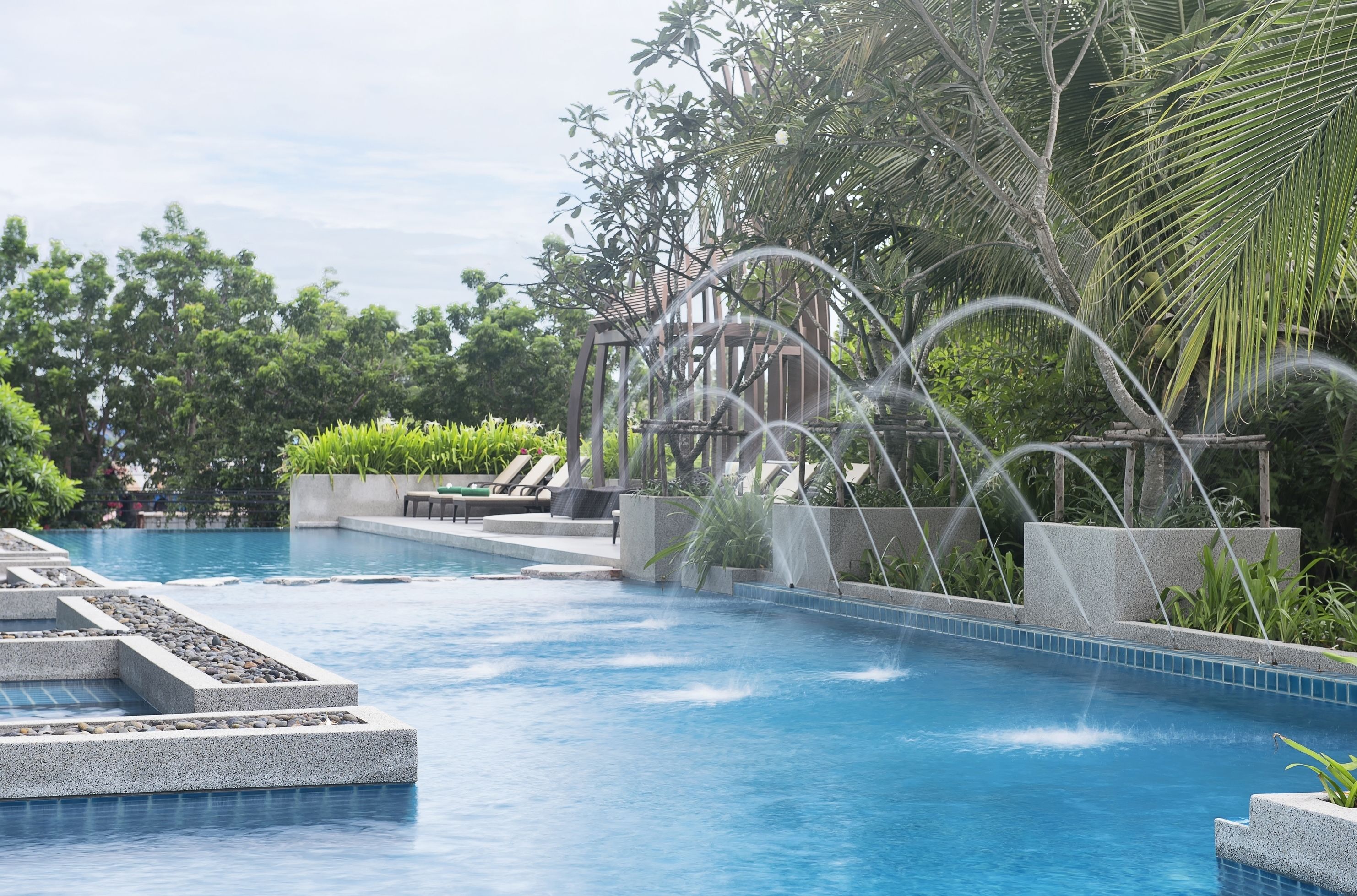 A swimming pool with a water feature, surrounded by greenery and a pergola structure.