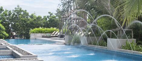 A swimming pool with a water feature, surrounded by greenery and a pergola structure.