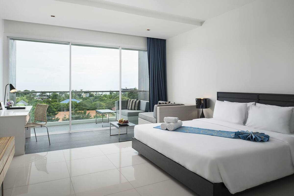 Habitación de hotel moderna con una cama grande, un balcón con vistas, un escritorio y una zona de estar.