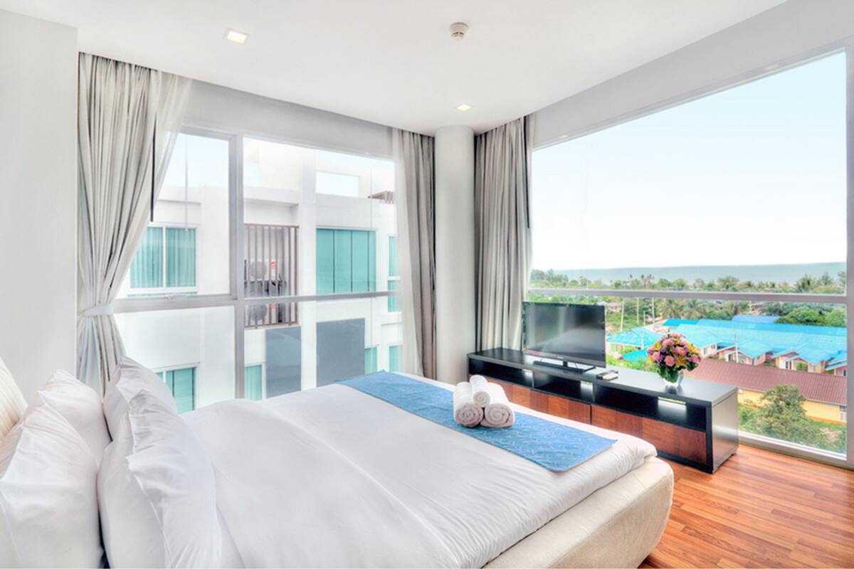 Una habitación de hotel moderna con una cama grande, un televisor sobre un mueble de madera, y un balcón con vistas a una piscina y edificios.