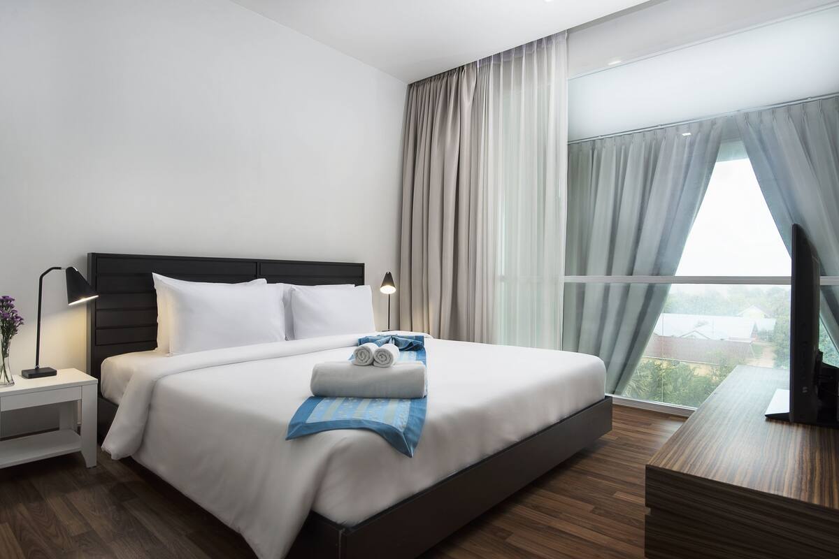 Una habitación de hotel moderna con una cama grande, una mesita de noche con lámpara, un escritorio de madera y vistas al exterior a través del ventanal.
