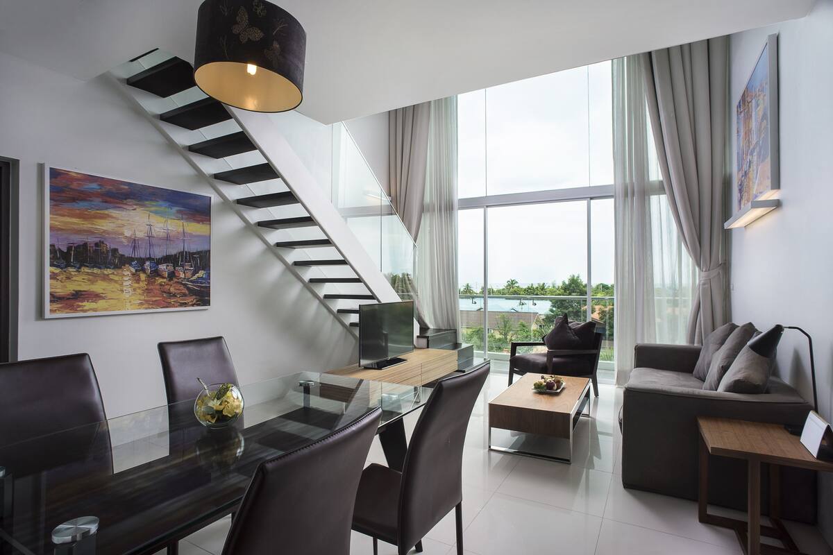 Una sala de estar moderna con una mesa de comedor de vidrio, asientos cómodos, una escalera y ventanas amplias que ofrecen vistas al exterior.