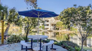 A patio with a table and benches, overlooking a calm lake and a building with balconies. - The Village At Palmetto Dunes (Hilton Head Island)
