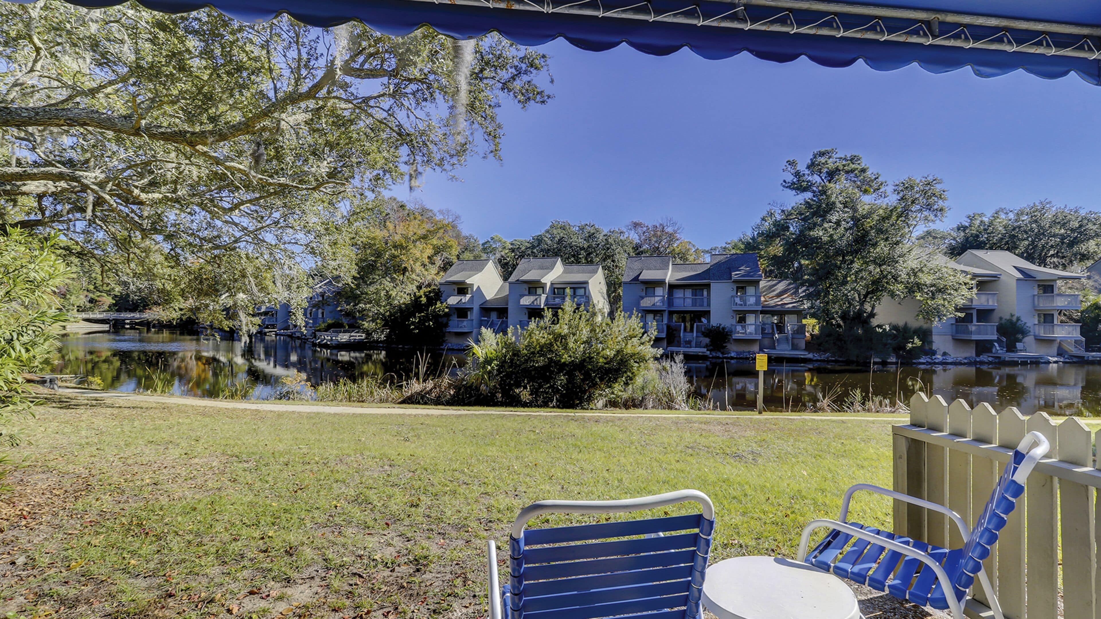 A view of a lakeside property with a covered patio area, outdoor seating, and a reflection of the buildings in the water.