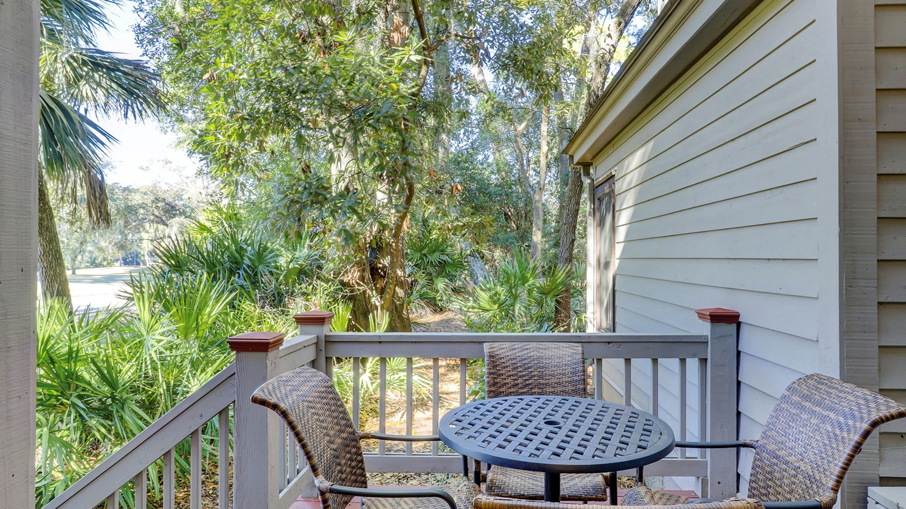 A covered patio with wicker furniture and a round table, surrounded by lush greenery and trees.