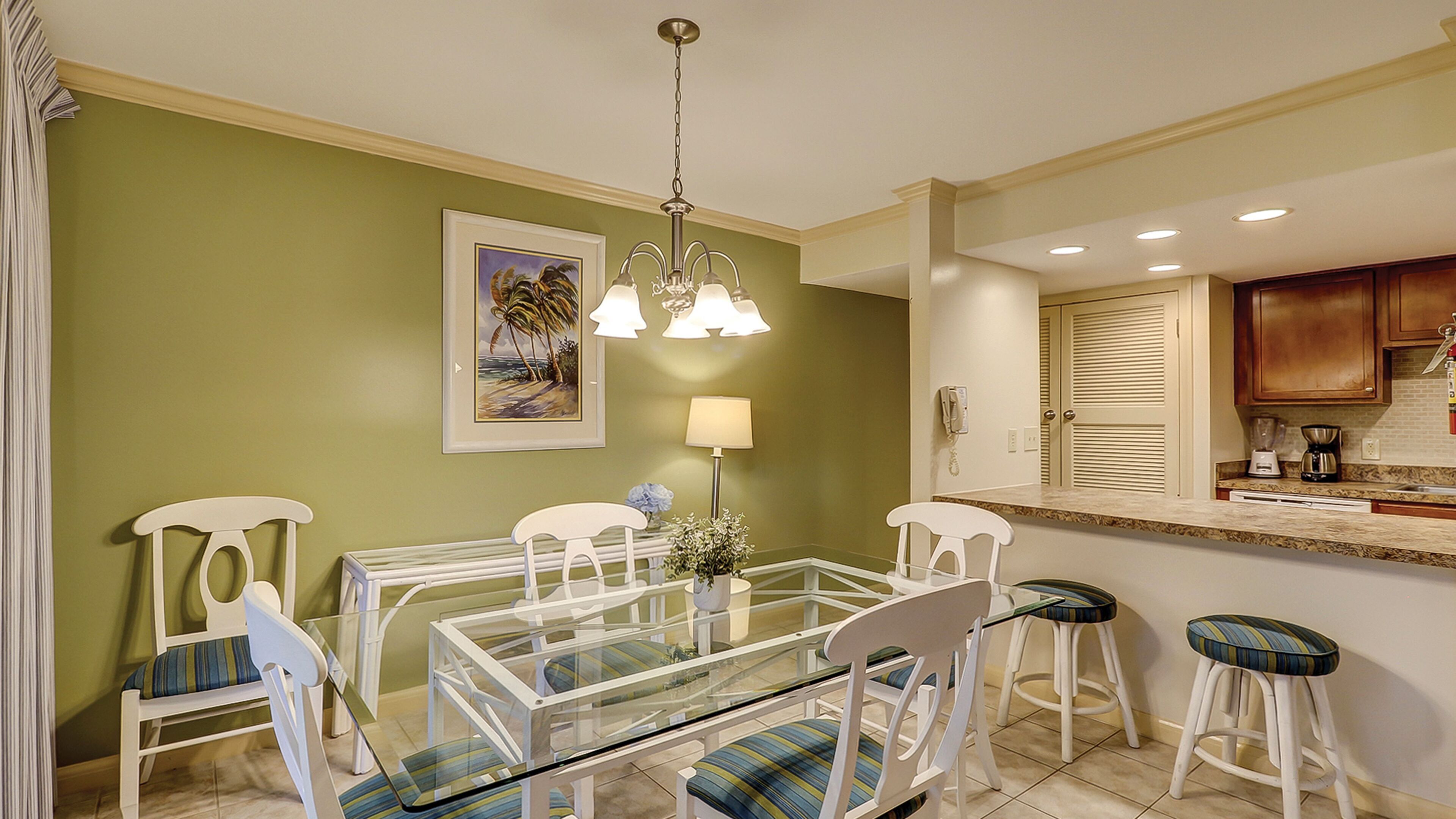 A dining area with a glass-top table, white chairs, a chandelier, a framed picture, and a kitchen in the background.