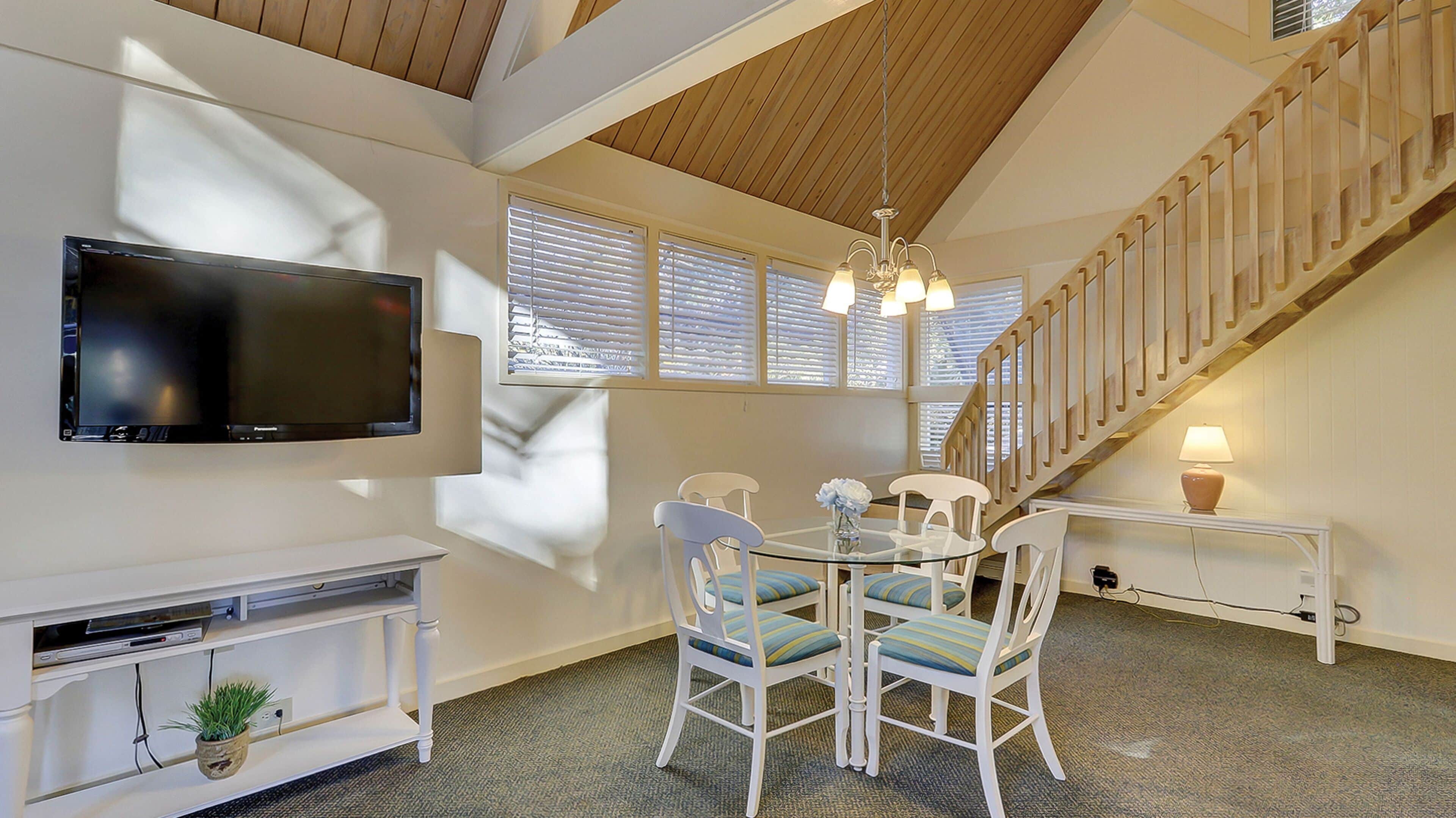 A dining area with a round table, white chairs, a television mounted on the wall, and a staircase with wooden railings.