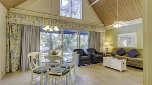 A spacious living area with a high ceiling, a dining table with chairs, a sofa, and a coffee table. Large windows with curtains provide a view of the outdoors. - The Village At Palmetto Dunes (Hilton Head Island)