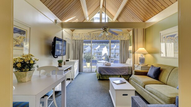 A hotel room with a bed, a desk, a chair, a television, a lamp, and a view of the outside. - The Village At Palmetto Dunes (Hilton Head Island)