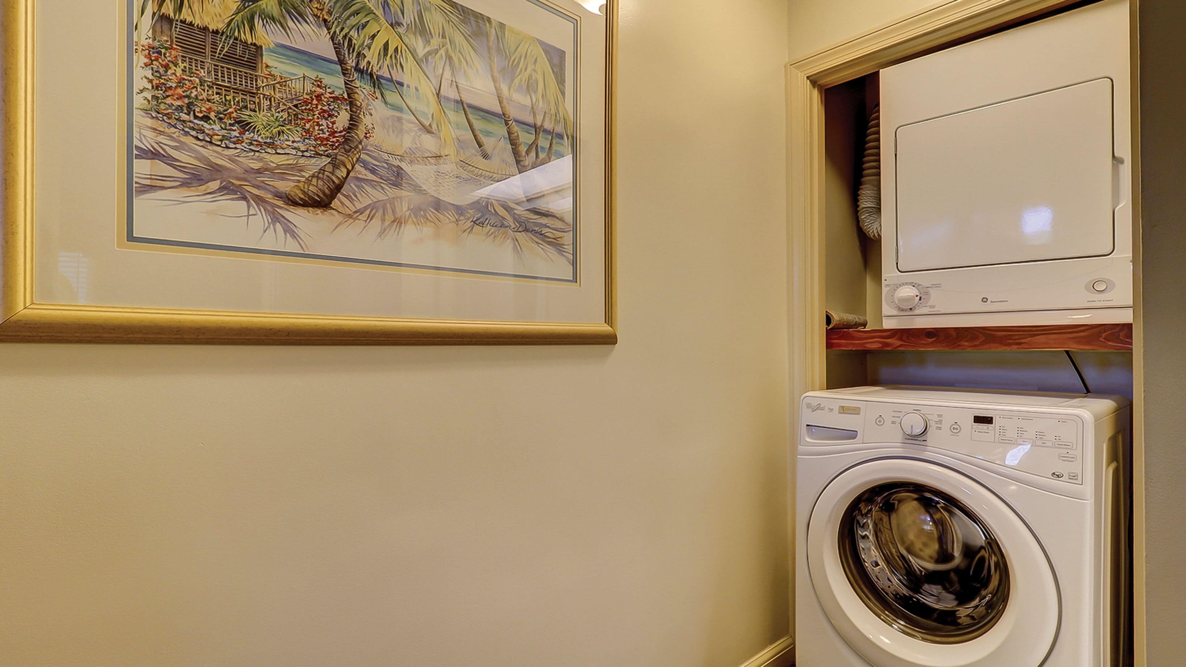 A laundry area with a washing machine and dryer, a framed tropical painting, and a wooden shelf.