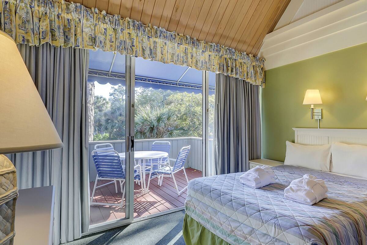 Een hotelkamer met een bed, twee bedlampjes, uitzicht op de omgeving en een balkon met stoelen en een tafel.