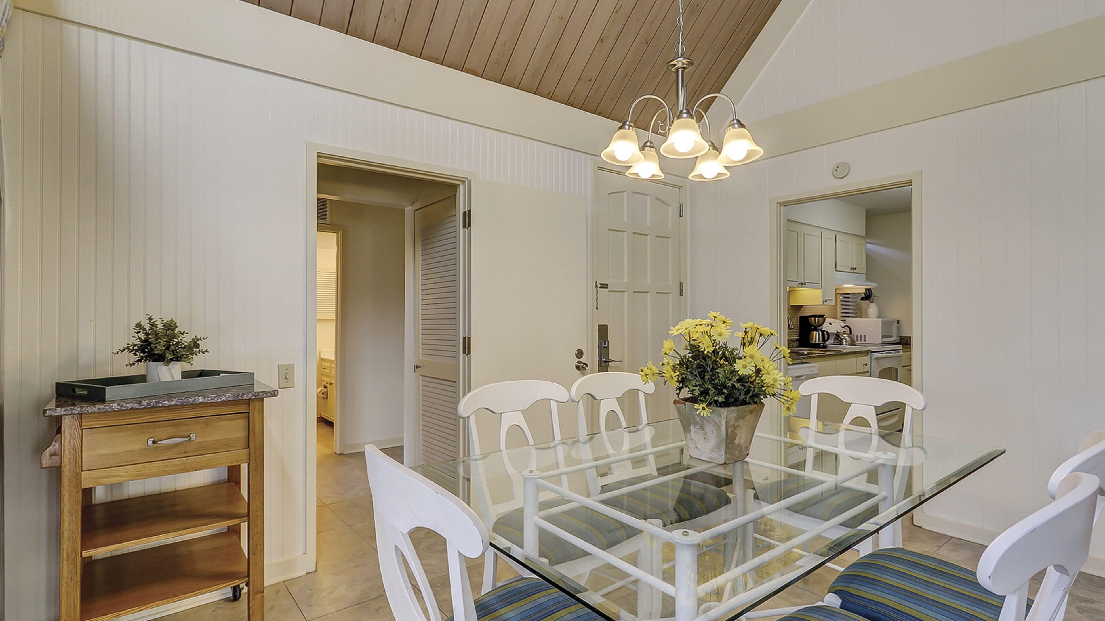 A dining area with a glass table surrounded by white chairs, a wooden cabinet, and a flower vase.