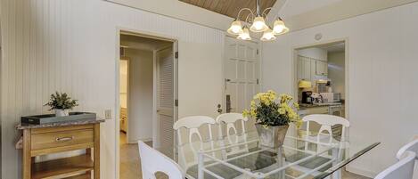 A dining area with a glass table surrounded by white chairs, a wooden cabinet, and a flower vase.