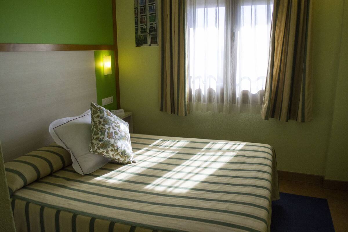 Una habitación de hotel con cama, almohada, mesita de noche, lámpara y ventana con cortinas.