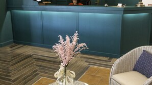 A reception area with a blue counter, a receptionist, a round wicker table with a vase of flowers, and two wicker chairs. - Hospedium Hotel Maestrazgo de Calatrava (Almagro)
