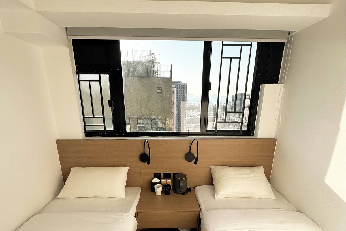 Ein kompaktes Hotelzimmer mit zwei Einzelbetten, einem kleinen Tisch mit Kaffeemaschine und einem Fenster mit Blick auf die Stadtlandschaft.